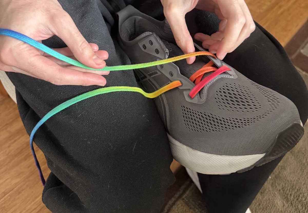 man threading rainbow-coloured shoelace through a shoe on his lap